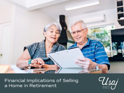 A retired couple sits at a glass table reviewing documents in their home. Below, white text on dark gray-blue background reads, "Financial Implications of Selling a Home in Retirement" next to the Usaj Realty logo.