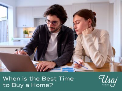 A couple dressed in business casual clothes sits at their kitchen table. They are reviewing documents and their laptop with concentration. Below the text reads "When is the Best Time to Buy a Home?" next to the Usaj Realty logo.