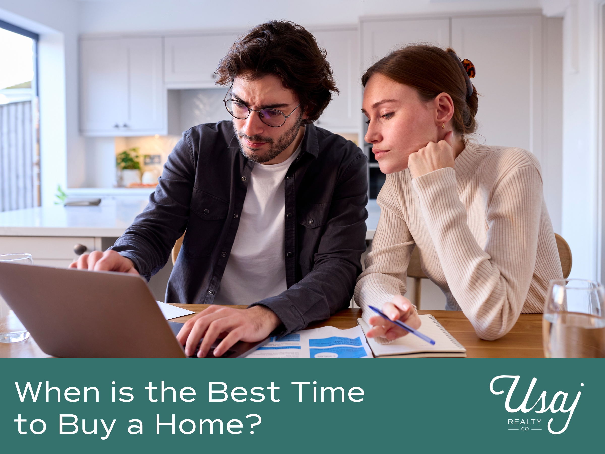 A couple dressed in business casual clothes sits at their kitchen table. They are reviewing documents and their laptop with concentration. Below the text reads "When is the Best Time to Buy a Home?" next to the Usaj Realty logo.