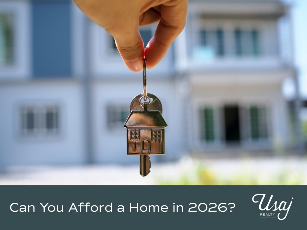 A photo of a person holding house keys dangles in the foreground, while a white house with blue trim is faded in the background. Below, white text on a charcoal background reads, "Can You Afford a Home in 2026?" next to the white Usaj Realty Logo.