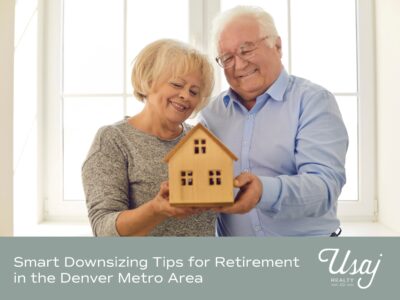 An elderly white couple holds a small wooden house in front of a plain white window. Below the white text and white Usaj Realty logo rest atop a gray background. The text reads: "Smart Downsizing Tips for Retirement in the Denver Metro Area"