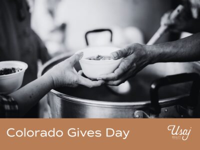 A black and white photo features someone dishing up a soup and handing it to another person. Below, white text on an orange background reads, "Colorado Gives Day" next to the Usaj Realty logo in white.