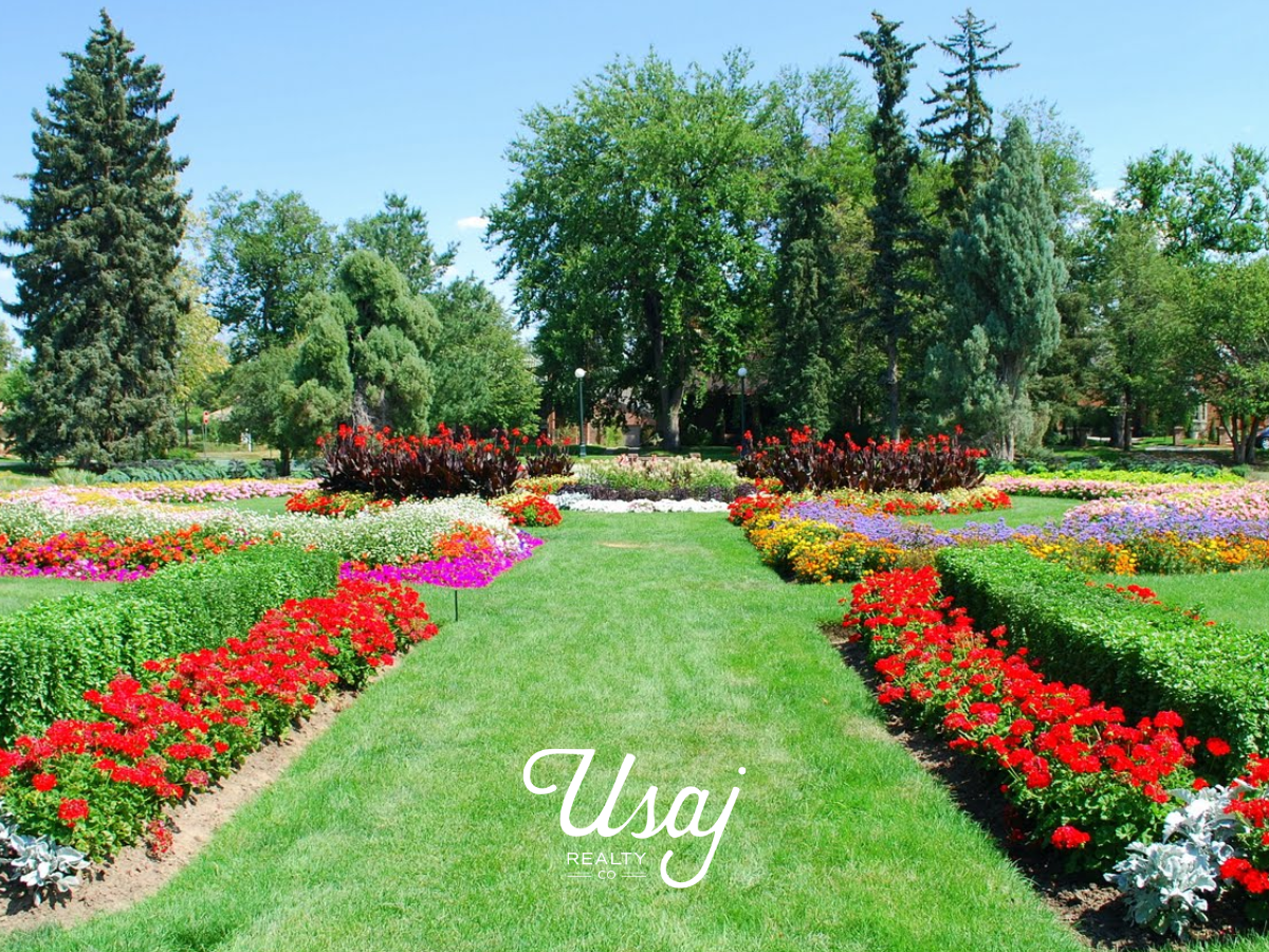 Red flowers and bushes line a grassy walkway in the Sunken Gardens in the Denver neighborhood Alamo Placita with the Usaj Realty logo in white at the center bottom of the photo.